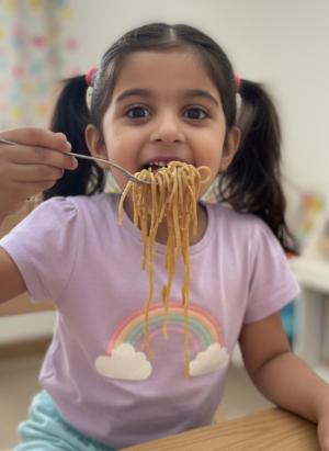 Young girl eating thi-thi noodles with a fork indoors, wearing a purple shirt with a rainbow design.